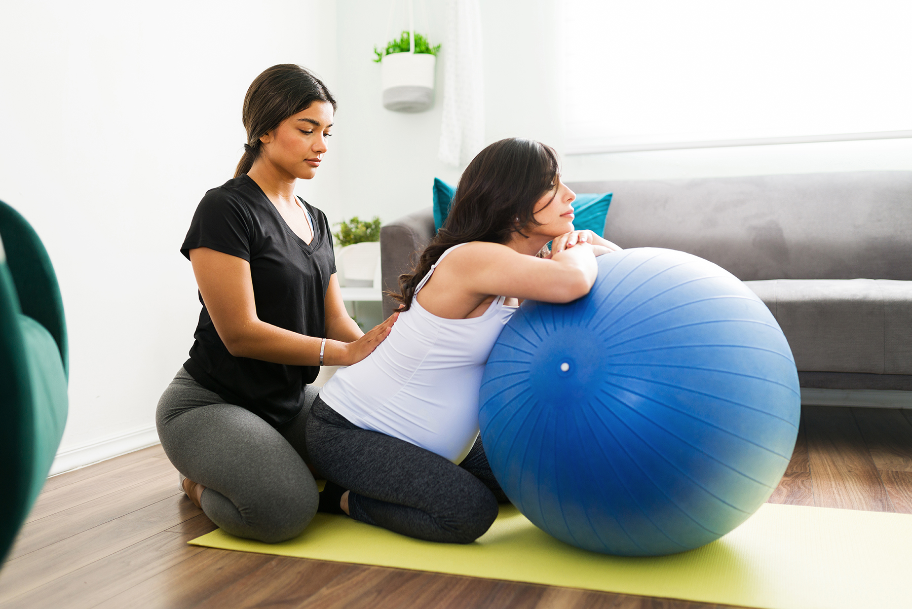 Beautiful doula in activewear helping a caucasian pregnant woman with her back pain. Expectant mother relaxing in a fitness ball while receiving a massage